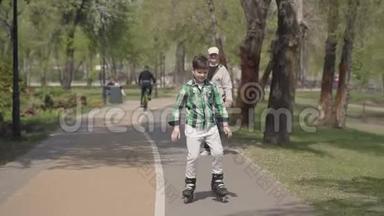 可爱的男孩穿着鲜艳的衣服在公园里滑旱冰，他的祖父走在附近。 户外休闲。 孩子玩得很开心